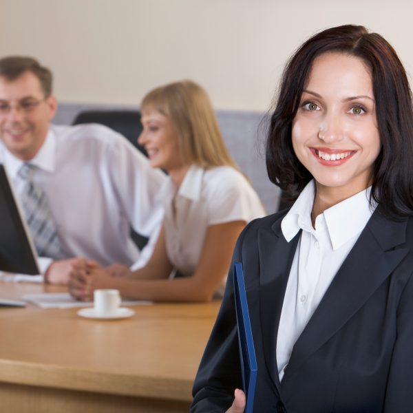 Portrait of brunette smiling businesswoman staring at camera holding blue paper case in her hands and two businesspeople on the background looking at the monitor